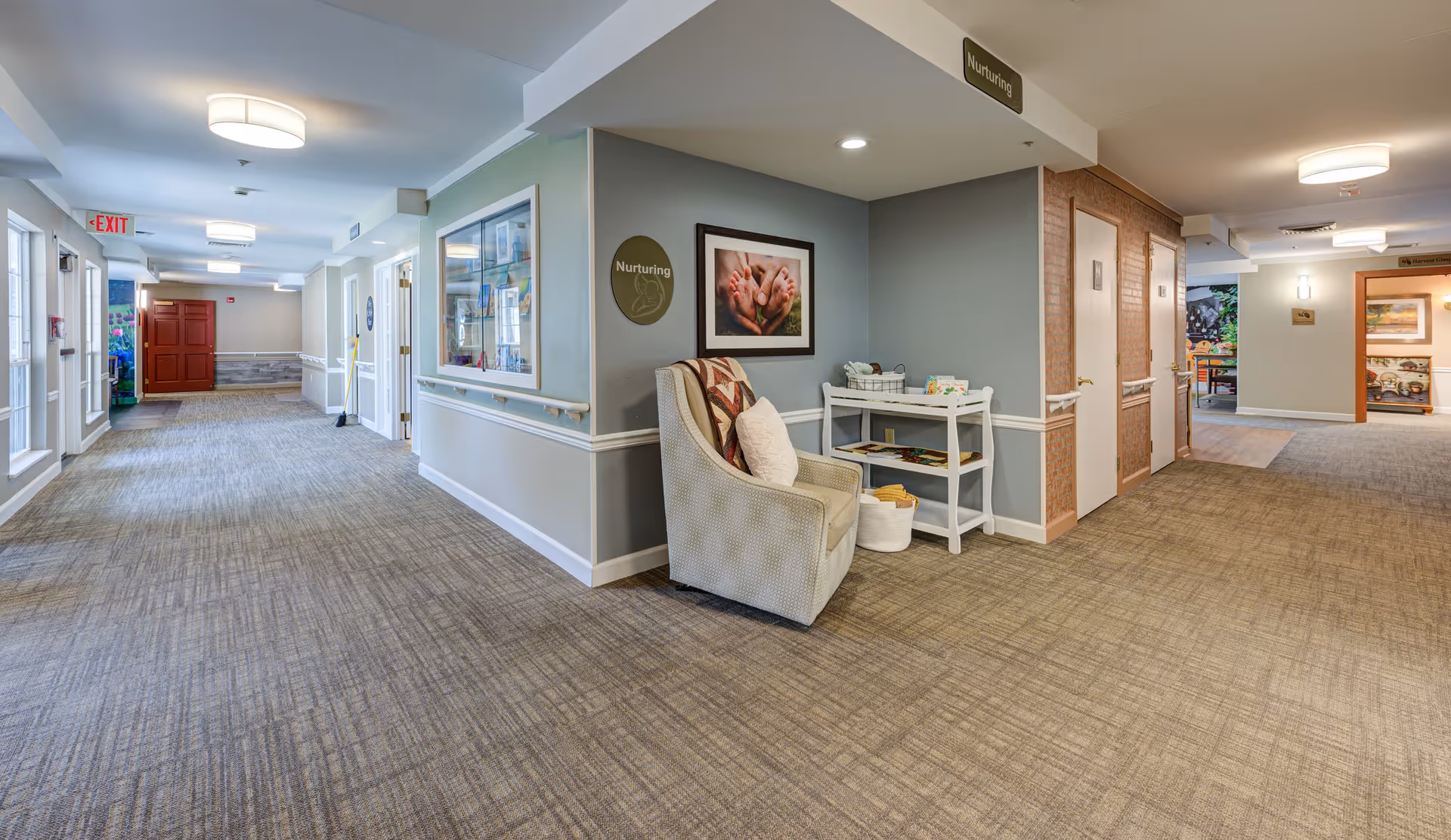 A wide hallway in a senior living facility with beige carpet and light gray walls. On the right side, there is a cozy armchair with a pillow and a small white table holding books and baskets. Above the chair is a framed photo of hands holding baby feet and a round sign that reads 'Nurturing.' Several doors line the hallway, and there are ceiling lights and an exit sign visible in the distance.