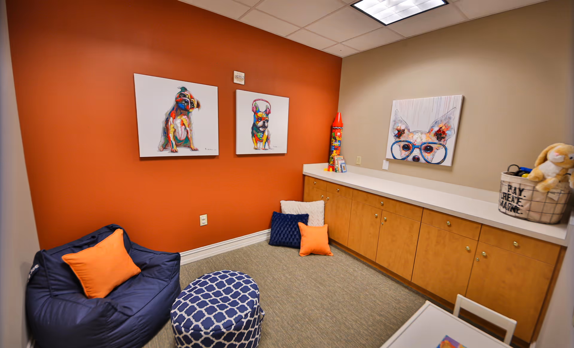 A cozy room with an orange accent wall featuring two colorful dog paintings. There is a navy blue bean bag chair with an orange pillow, a blue patterned ottoman, and several cushions on the floor. A long wooden cabinet with a white countertop lines the opposite wall, decorated with a large painting of a dog wearing glasses, a tall red toy rocket, and a basket with a stuffed bunny and the words 'PLAY CREATE IMAGINE'.