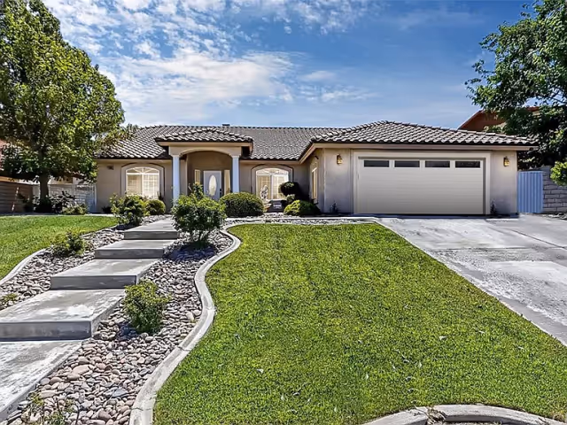 Front exterior view of a single-story house with a tiled roof, a double garage door, a concrete driveway, a curved walkway with steps leading to the front door, and a well-maintained lawn with shrubs and trees under a partly cloudy sky.