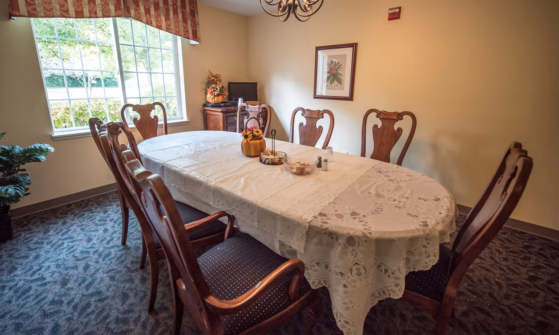 Small dining room with an oval table covered by a lace tablecloth surrounded by wooden chairs and a window letting in daylight.