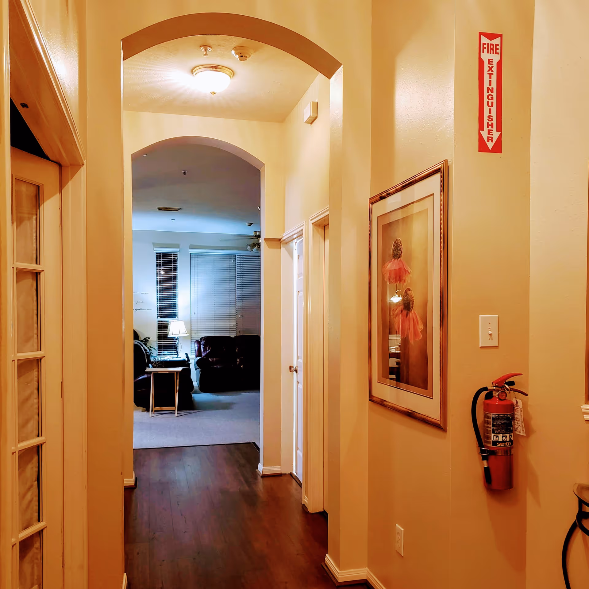 Interior hallway with wooden floor leading to a living room area. The hallway has beige walls, a framed picture on the right wall, and a fire extinguisher mounted below a fire extinguisher sign. The living room visible at the end has a lamp on a side table, a couch, and windows with blinds.