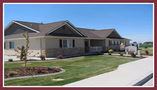 Exterior view of a single-story building with beige siding and stone accents, surrounded by a well-maintained lawn and landscaping. A sidewalk runs along the front, and a sign near the entrance reads Ashley Manor Memory Care.