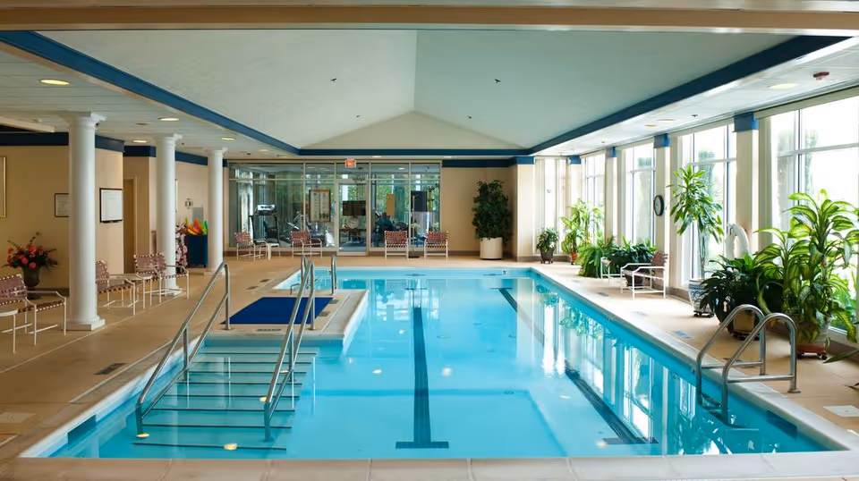 Indoor swimming pool area with clear blue water, metal handrails, and steps leading into the pool. The room has large windows on one side with several green plants placed along the windowsill. There are multiple chairs and seating areas around the pool, with white columns and a high ceiling.