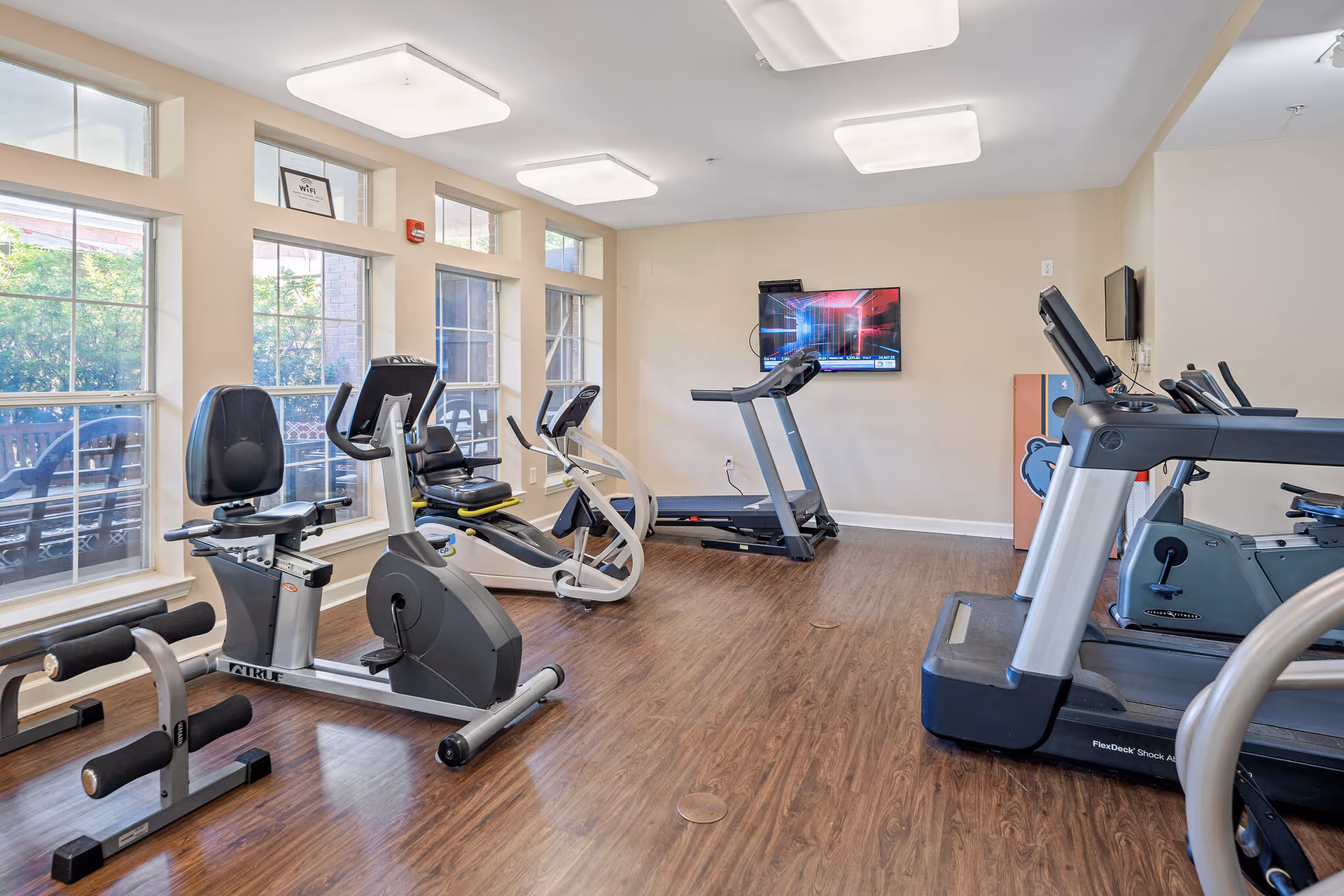 A fitness room with exercise equipment including stationary bikes, treadmills, and a rowing machine. The room has large windows letting in natural light, wood flooring, and a wall-mounted TV.