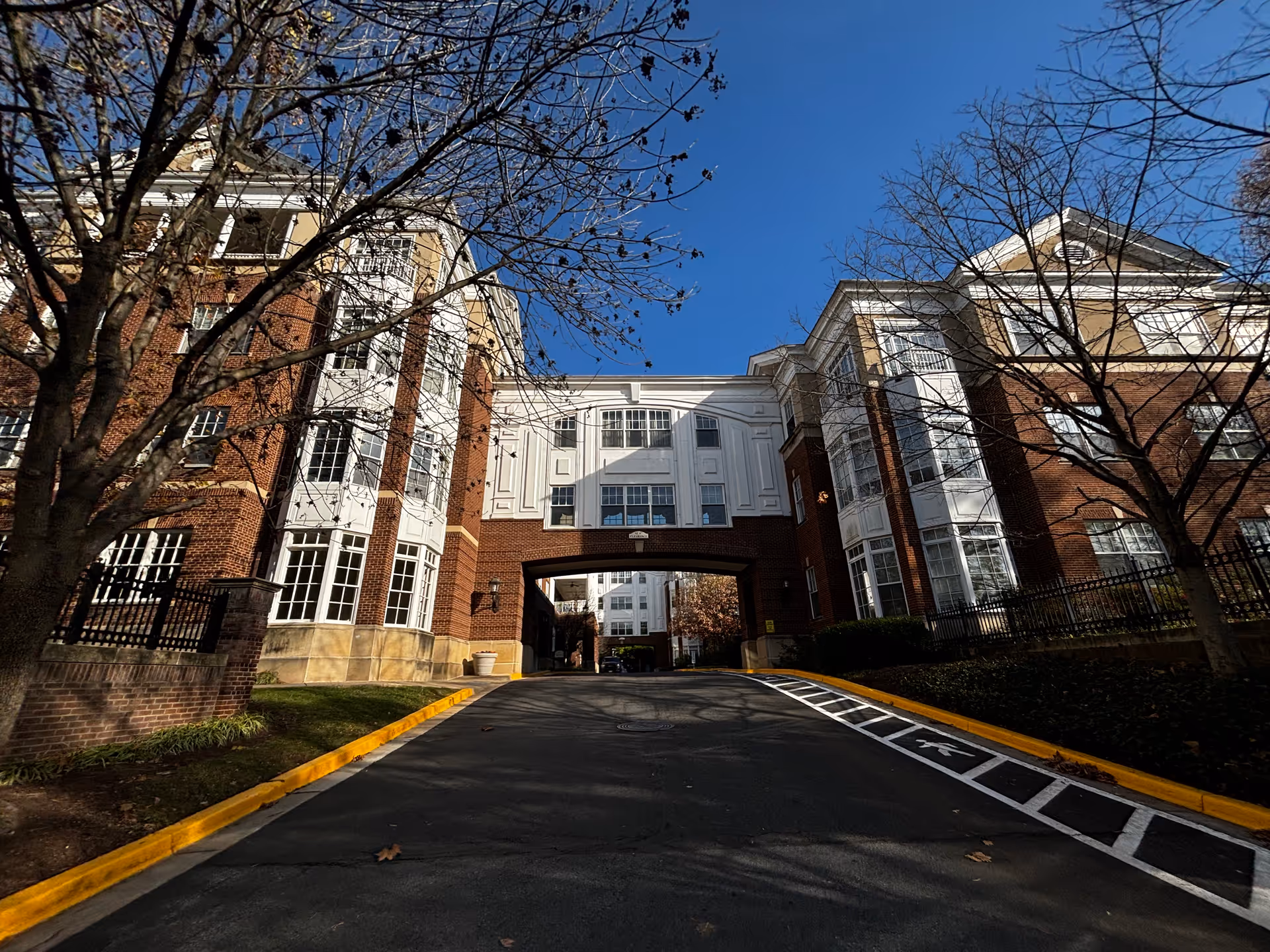 Exterior view of a multi-story brick and white-paneled building with large windows, an archway over a driveway, and leafless trees on either side under a clear blue sky.