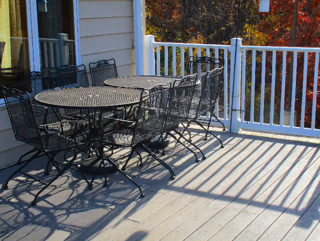 Outdoor patio area with a black metal round table and six matching chairs on a wooden deck. The deck has white railing and is adjacent to a building with beige siding. Trees with autumn-colored leaves are visible in the background.
