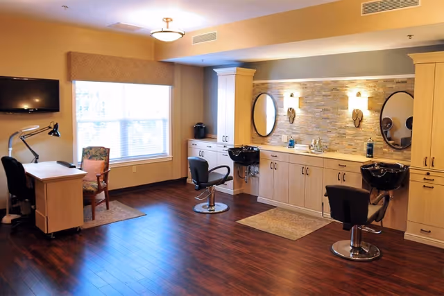 A bright salon-style interior with two shampoo stations and styling chairs, mirrors, a desk, and a large window.