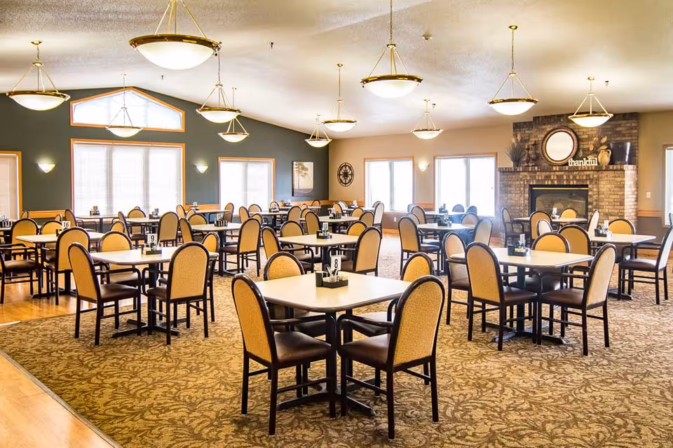 A spacious dining room with multiple square tables, each surrounded by four chairs with beige upholstery and dark wooden frames. The room features large windows allowing natural light, a carpeted floor with a patterned design, and a brick fireplace with a round mirror and decorative items on the mantel. Several hanging light fixtures illuminate the area.