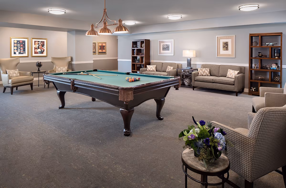 A spacious senior living common area featuring a pool table with billiard balls and cues arranged for a game. The room is furnished with comfortable armchairs, sofas, side tables, bookshelves, and decorative framed artwork on the walls. A small round table with a floral arrangement is in the foreground.