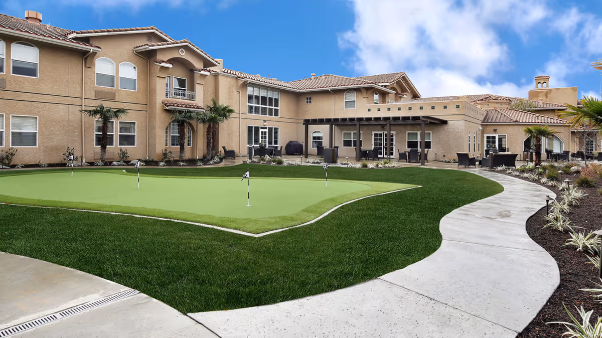 Courtyard of a beige senior living facility with a putting green, curved concrete walkway, patio seating, and palm trees.