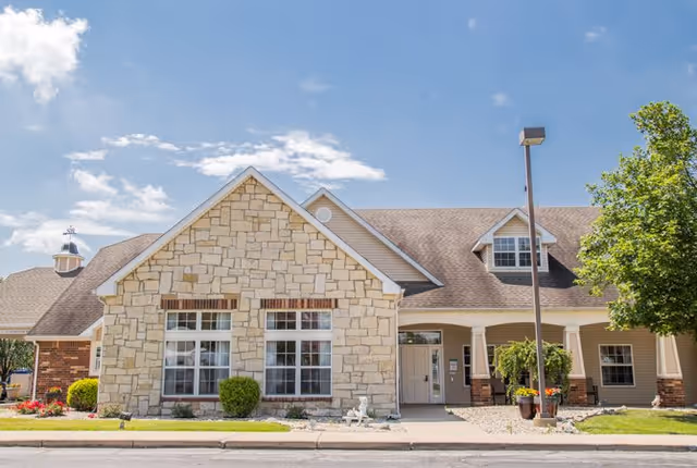 Exterior front view of a single-story building with a stone facade and beige siding, featuring multiple windows, a covered entrance, a lamp post, and landscaping with bushes and flowers under a partly cloudy blue sky.