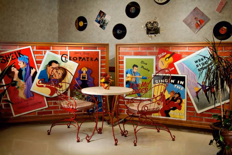 Retro-themed seating area with a round table, two red wrought-iron chairs, and colorful vintage posters and vinyl records on the walls.
