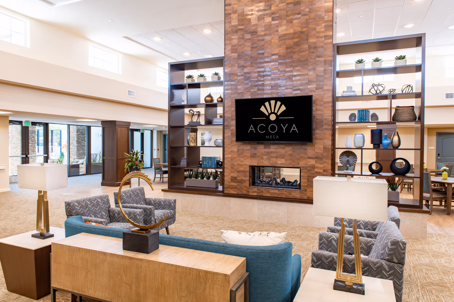 A spacious and well-lit living room area in Acoya Mesa featuring modern furniture including a blue sofa and patterned armchairs arranged around a central fireplace with a large TV above it displaying the Acoya Mesa logo. The room has tall shelves with decorative items and large windows allowing natural light to fill the space.