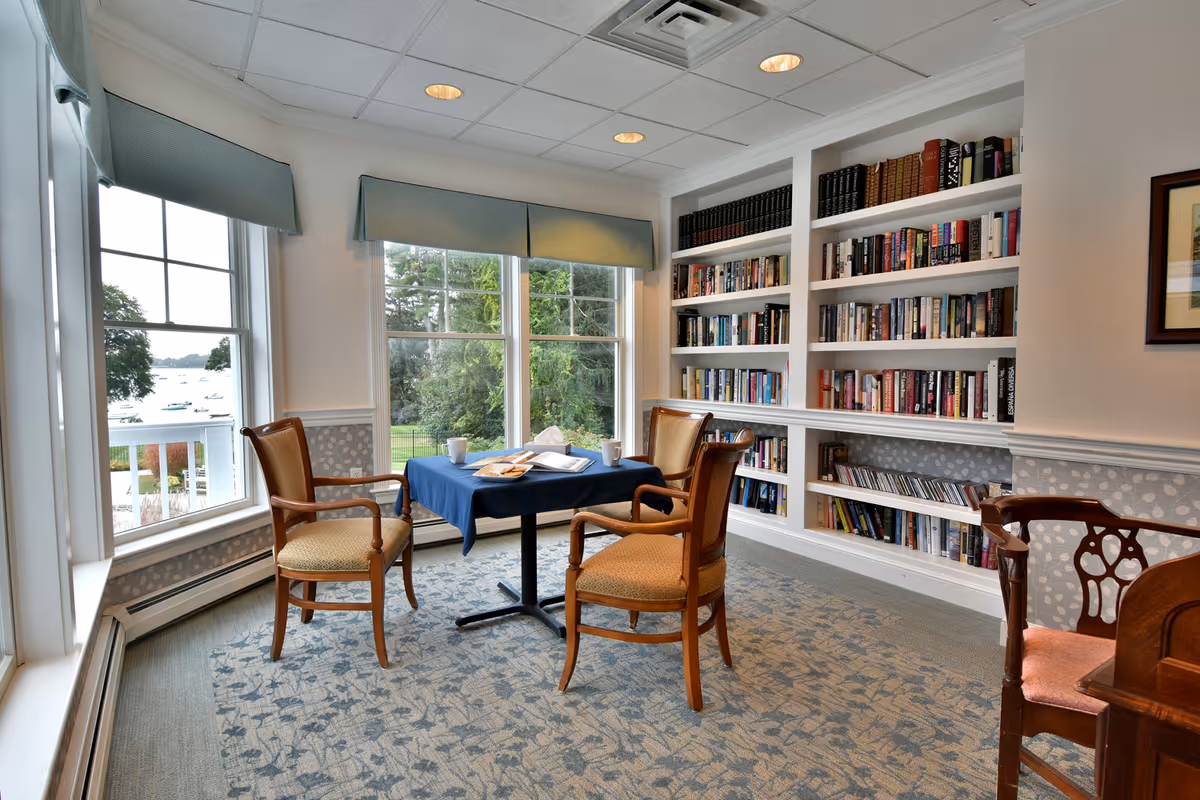 Bright reading room with built-in bookshelves, a table with a blue cloth and wooden chairs by large windows overlooking trees and water.