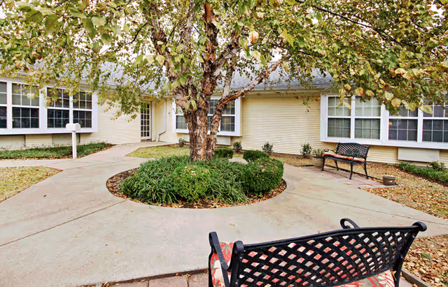 Outdoor courtyard area at American House Hendersonville featuring a circular concrete walkway surrounding a tree with green shrubs. There are two black metal benches with patterned cushions placed along the walkway. The building exterior is light-colored with multiple windows.