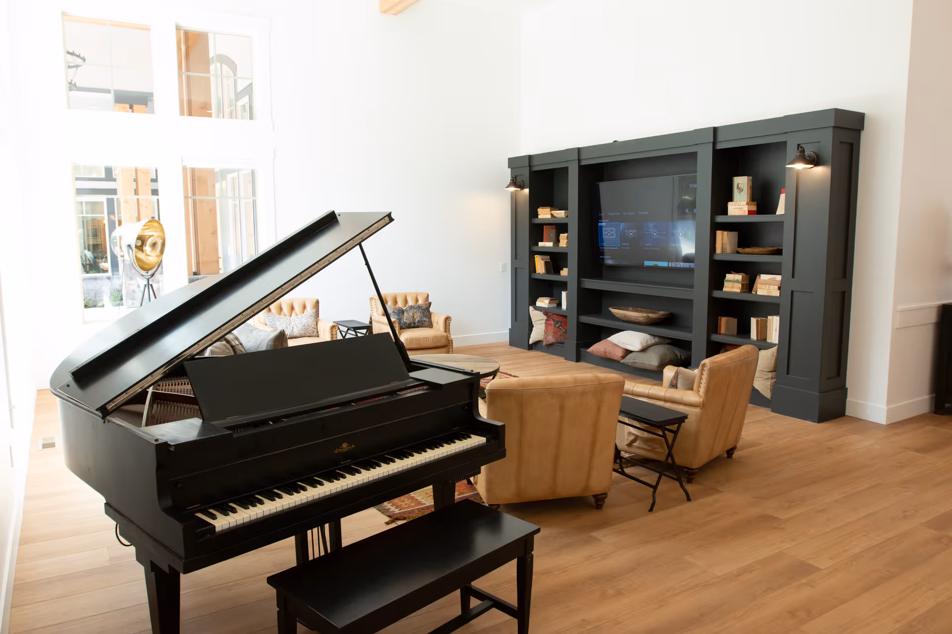 A bright and spacious living room featuring a black grand piano in the foreground, tan leather armchairs arranged around a low wooden coffee table, and a large dark built-in shelving unit with a flat-screen TV and decorative books and pillows. Large windows allow natural light to fill the room, and a vintage-style spotlight lamp is visible near the windows.