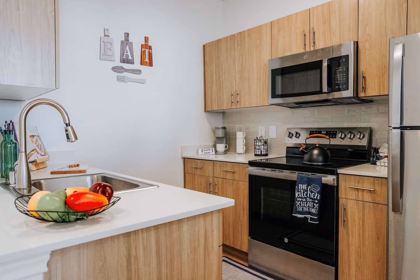 Modern kitchen with wooden cabinets, stainless steel appliances including a microwave, stove, and refrigerator. A white countertop with a sink and a bowl of colorful artificial fruits is visible. Wall decor spells out the word 'EAT' with cutlery shapes underneath.