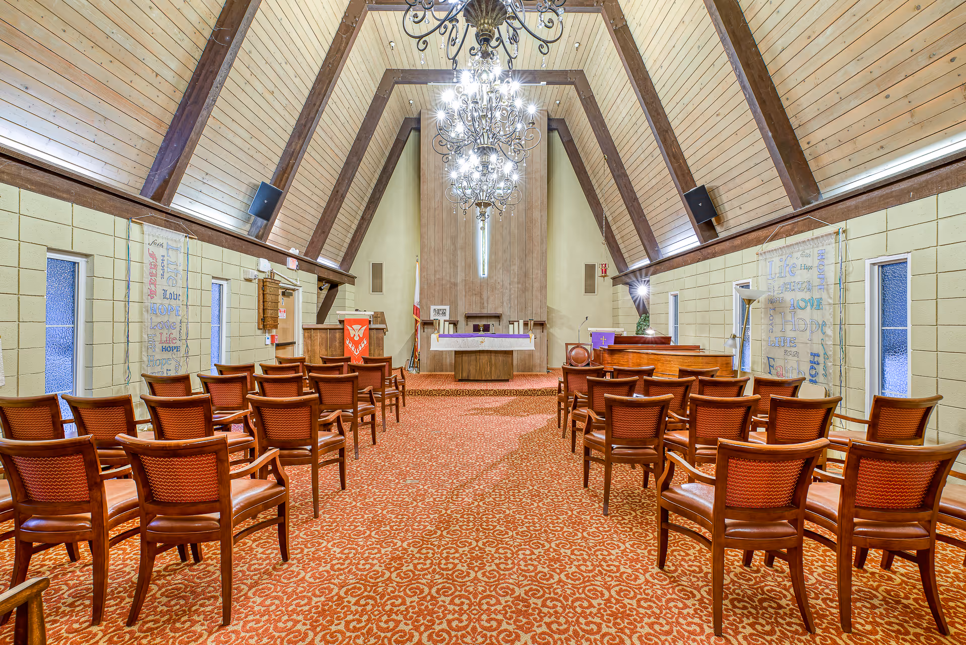 Interior view of a chapel with wooden A-frame ceiling, chandeliers, rows of wooden chairs with red cushions, a red patterned carpet, and an altar at the front with religious banners on the walls.