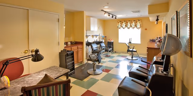 Interior view of a salon room in a senior living facility with salon chairs, hair dryers, a table with a lamp, and cabinets. The room has a checkered floor with green, white, and red tiles, yellow walls, and a window with floral curtains letting in natural light.