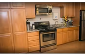 A kitchen area with wooden cabinets, a stainless steel stove with oven, a microwave above the stove, a stainless steel refrigerator, a granite countertop with a sink, and a coffee maker on the counter. There is a small vase with yellow flowers on the countertop.