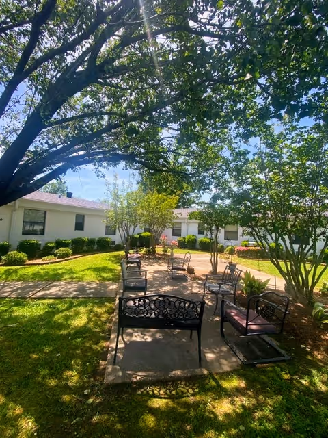 Shaded courtyard with metal benches and chairs under large trees in front of a single-story white building.