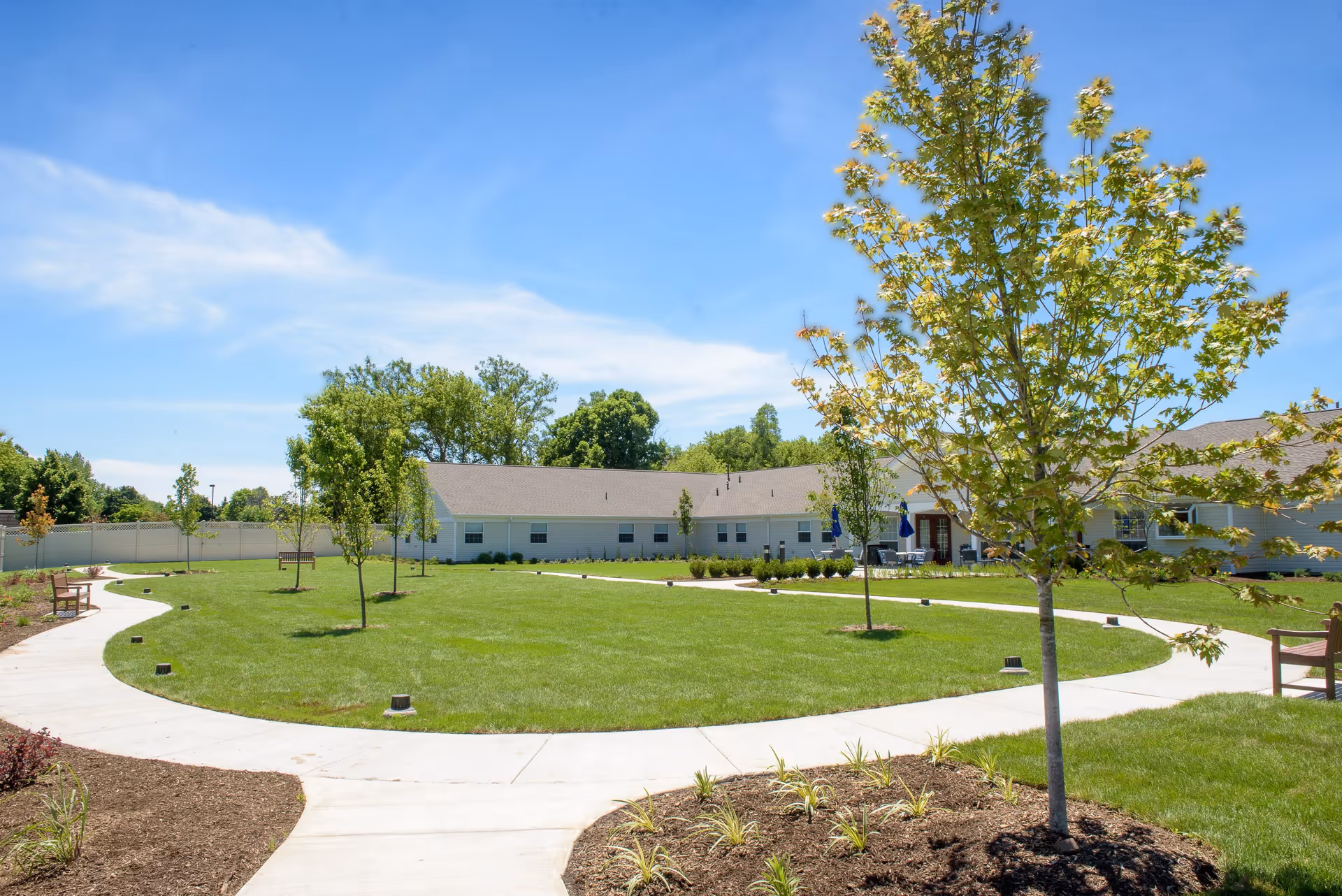 A sunny outdoor courtyard with a circular concrete walkway surrounding a grassy area with young trees and benches. Single-story buildings with light-colored siding and a gray roof are visible in the background under a clear blue sky.