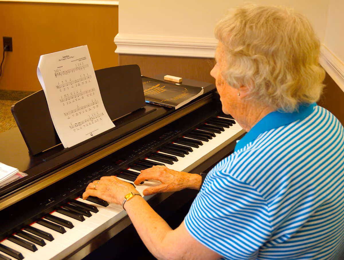 An elderly woman with short curly hair wearing a blue and white striped shirt is playing a digital piano. Sheet music titled 'America (My Country 'Tis of Thee)' is placed on the piano stand. The setting appears to be an indoor room with beige walls and a carpeted floor.