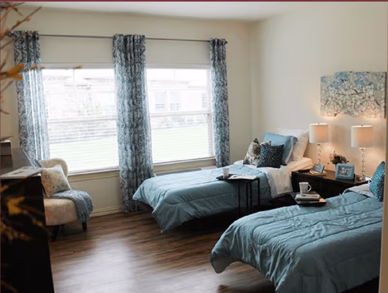 A bright bedroom with two twin beds dressed in light blue bedding and multiple pillows. Between the beds is a dark wooden nightstand with two lamps, a small plant, and framed photos. A large window with patterned blue curtains lets in natural light. There is a cushioned chair with a throw blanket and pillow near the window. The floor is wood, and the walls are painted light beige.