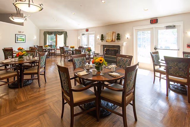 A bright dining room with several round wooden tables set with white cups, napkins, and floral centerpieces. The room features wooden flooring, a fireplace with decorative items on the mantel, large windows with green curtains, and glass doors leading outside. The space is well-lit with ceiling lights and hanging light fixtures.