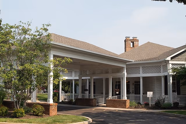 Front entrance of a single-story senior living building with a covered porte-cochere supported by white columns and brick planters.