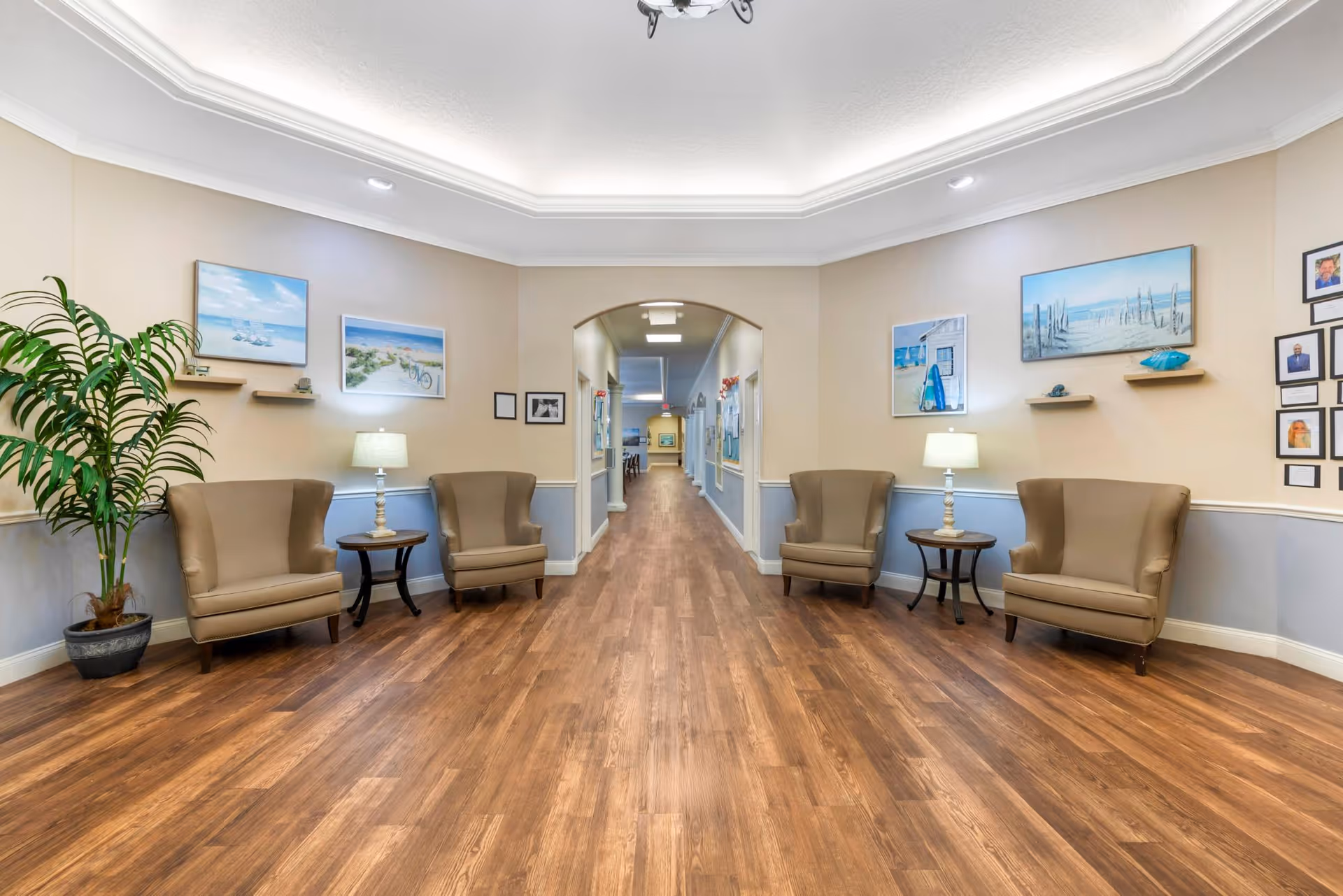 A wide hallway in a senior living facility with wooden flooring, beige walls with blue wainscoting, and a white ceiling with recessed lighting. Four beige armchairs are arranged in pairs on either side of the hallway, each pair separated by a small round table with a lamp. The walls are decorated with framed beach-themed artwork and photographs. A large potted plant is placed on the left side near the chairs. The hallway extends into the distance with more artwork and lighting visible.