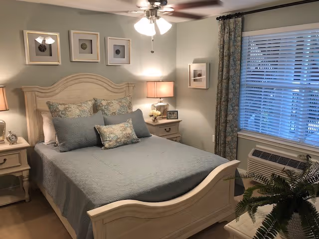 A cozy bedroom with a wooden bed frame and headboard, neatly made with a blue quilt and multiple decorative pillows. There are two matching wooden nightstands on either side of the bed, each with a lamp. Above the bed are three framed pictures. A window with blinds and patterned curtains is on the right wall, and a ceiling fan with lights is overhead. A potted plant is visible in the foreground.