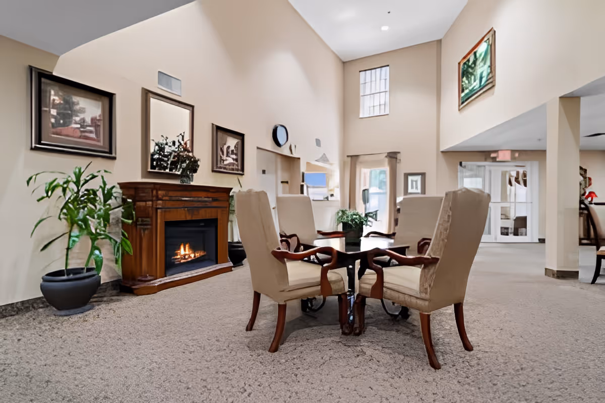 A cozy sitting area in a senior living facility with four beige upholstered chairs around a wooden table. There is a lit fireplace with a wooden mantle, potted plants, and framed artwork on the walls. The room has high ceilings and large windows allowing natural light to enter.