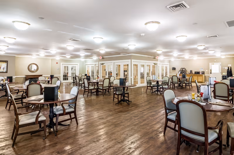 Large, well-lit dining room with multiple round tables and upholstered chairs on hardwood floors.
