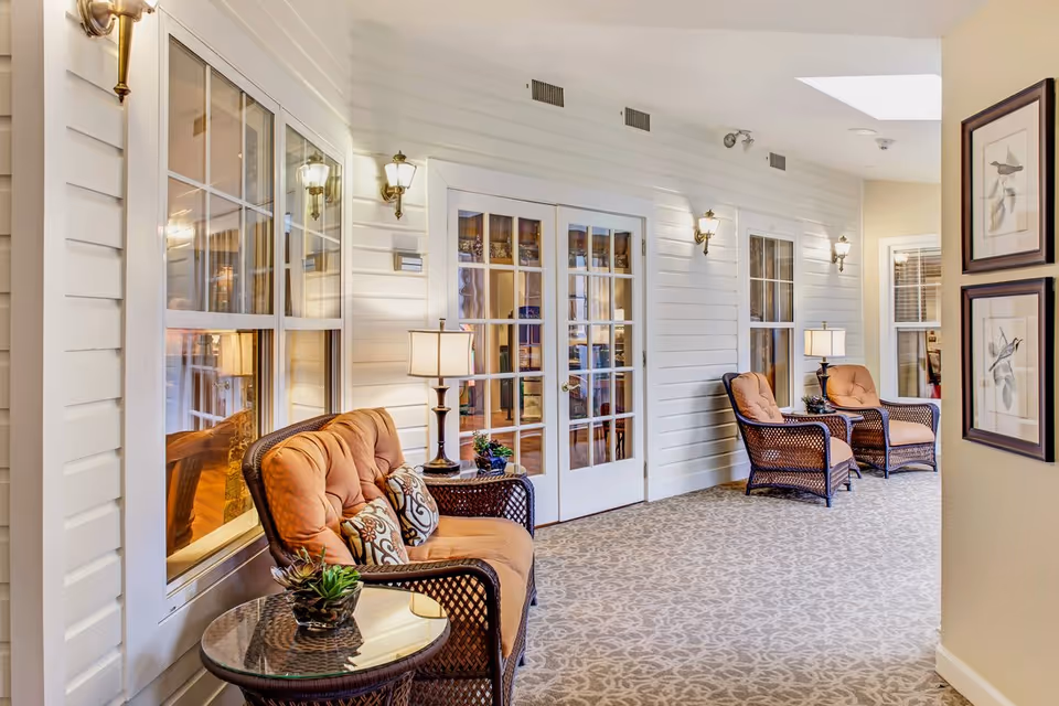 A bright indoor sitting area with wicker chairs, side tables, and lamps along a wall of windows and glass double doors.