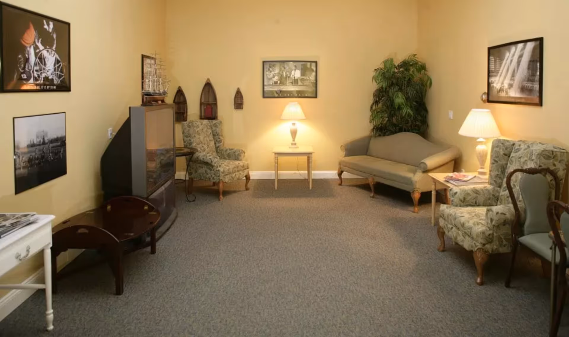 A cozy living room with beige walls and carpeted floor, featuring a vintage-style TV on a stand, two floral armchairs, a green sofa, two side tables with lamps, a potted plant, and framed pictures on the walls.