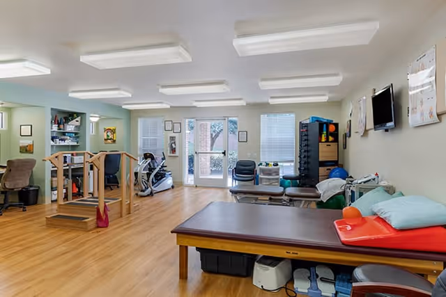 A bright therapy room with treatment tables, exercise equipment, parallel bars, and storage cabinets.