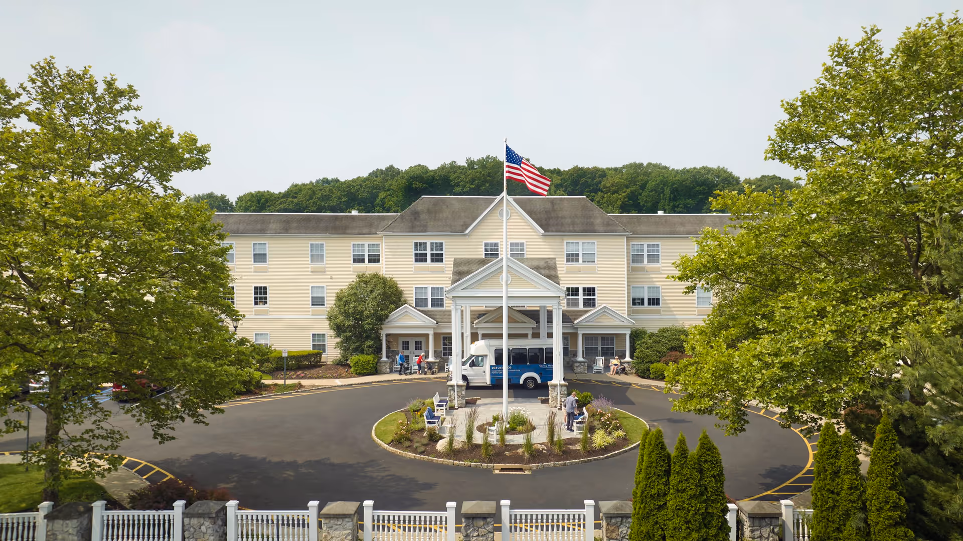 Front exterior view of Monarch Spring Meadows facility with a circular driveway, an American flag on a flagpole in the center, surrounded by benches and landscaping. The building is three stories tall with multiple windows, and there are trees and greenery around the property.