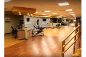 Spacious indoor fitness room with stationary bikes and other exercise equipment on wood floors, a reception desk area, and overhead lighting.