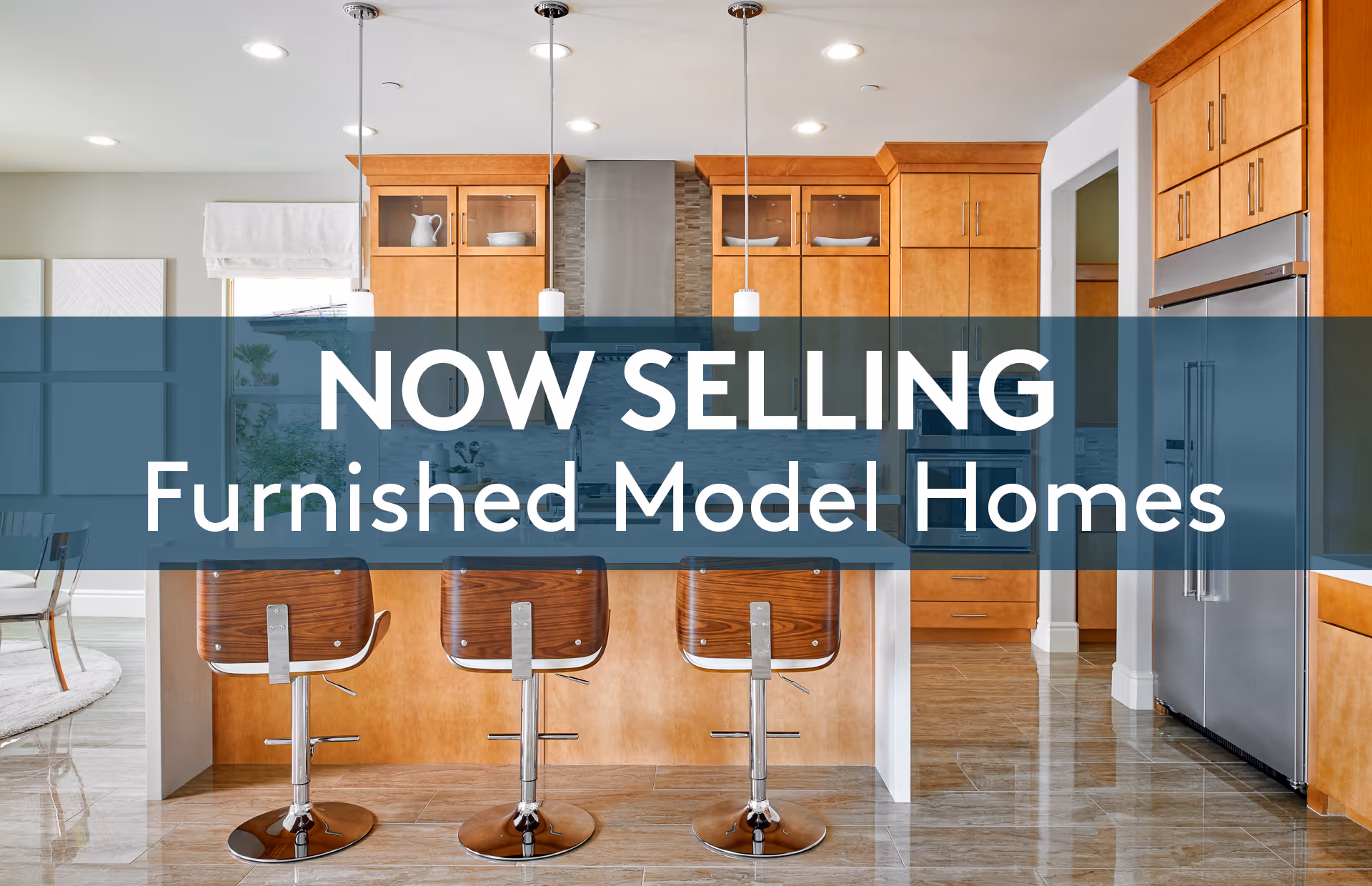 Modern kitchen with wooden cabinets, stainless steel appliances, and three wooden bar stools at a kitchen island. The kitchen is well-lit with pendant lights and has a tiled floor. A dining area is partially visible to the left.