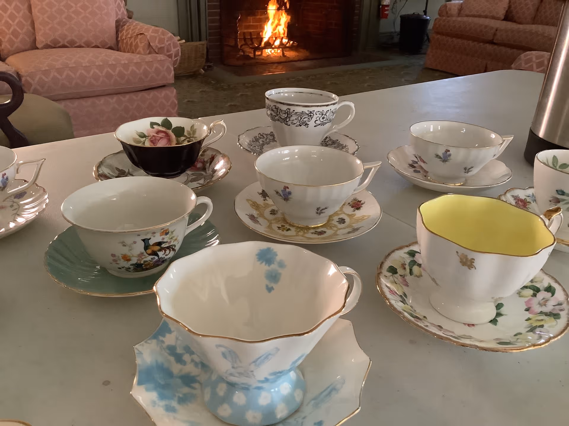 A collection of assorted vintage teacups and saucers arranged on a white table in a cozy room with a lit fireplace and pink upholstered chairs in the background.