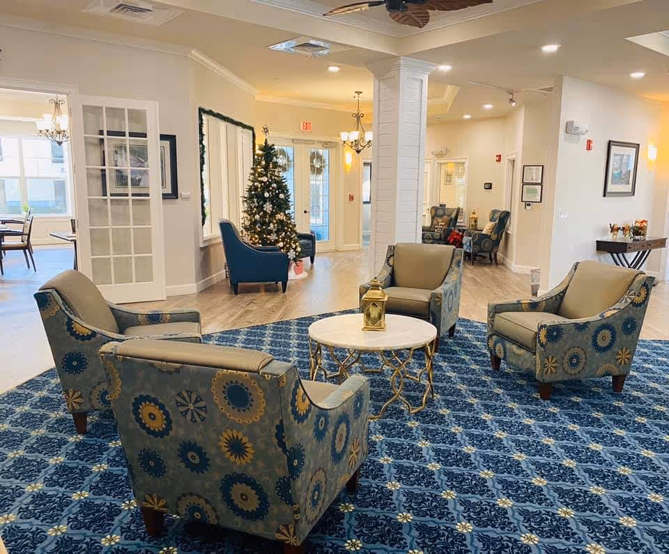 A cozy senior living facility common area with four patterned armchairs arranged around a small round table with a decorative lantern on it. The room features a blue patterned carpet, light-colored walls, and wood flooring. In the background, there is a decorated Christmas tree, a large mirror, and additional seating. The space is well-lit with ceiling lights and chandeliers, and there is a doorway leading to another room with dining tables and chairs.