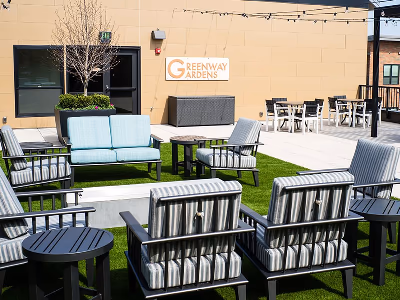 Outdoor rooftop patio with cushioned chairs and tables on artificial turf in front of a building wall displaying a 'Greenway Gardens' sign.