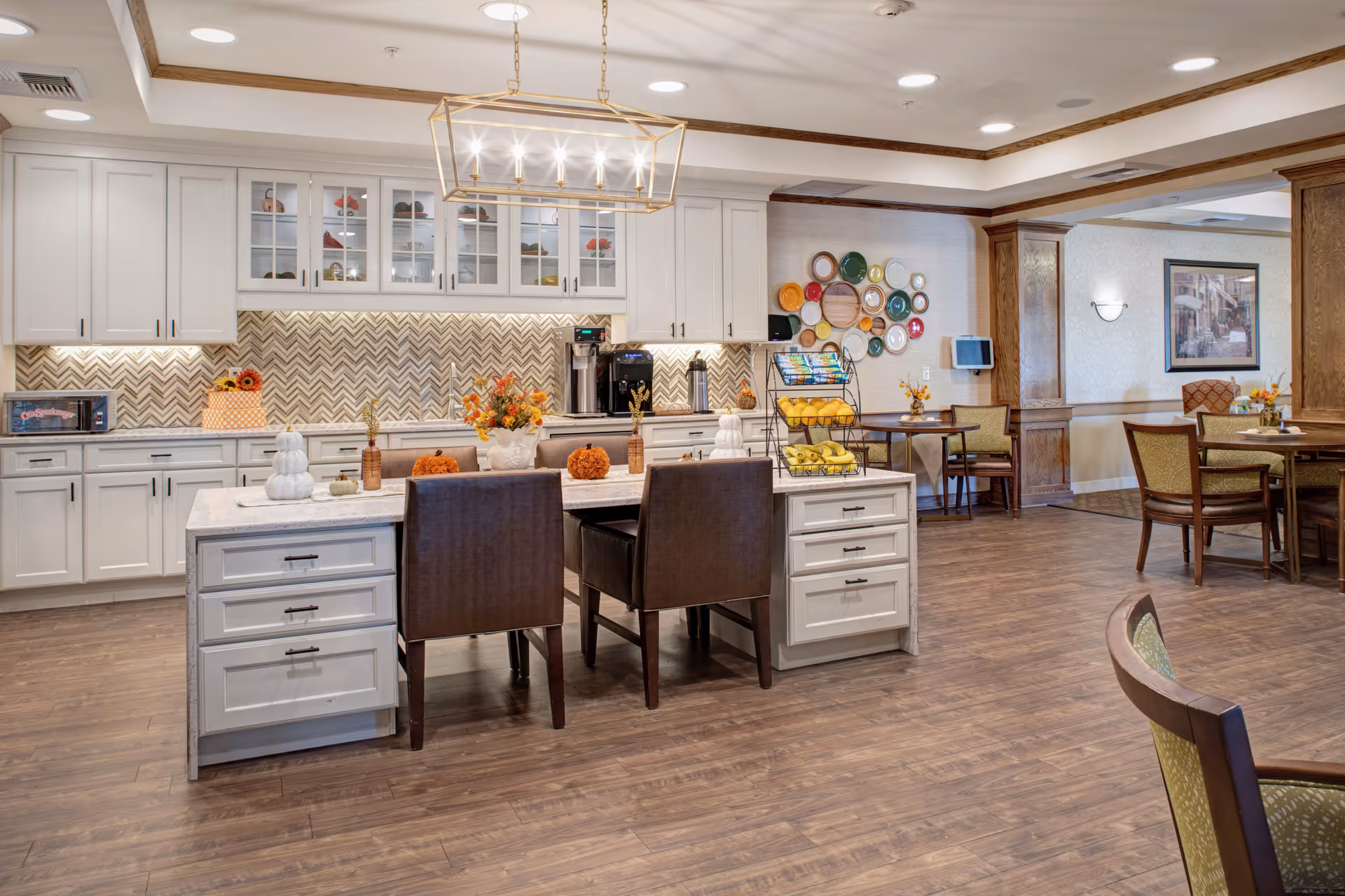 Bright communal kitchen and dining area with a large island, white cabinetry, seating and decorative plates on the wall.