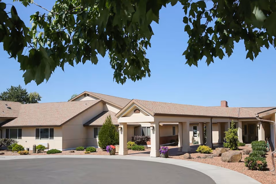 Exterior view of a single-story beige building with a covered entrance, surrounded by landscaping including bushes, rocks, and potted flowers, under a clear blue sky with tree leaves partially visible at the top.