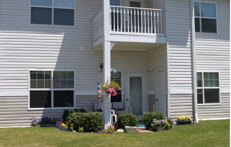Exterior view of a two-story building with white siding, featuring a small patio area with chairs, a hanging flower basket, and various flowering plants and shrubs around the patio. There are multiple windows and a balcony above the patio.