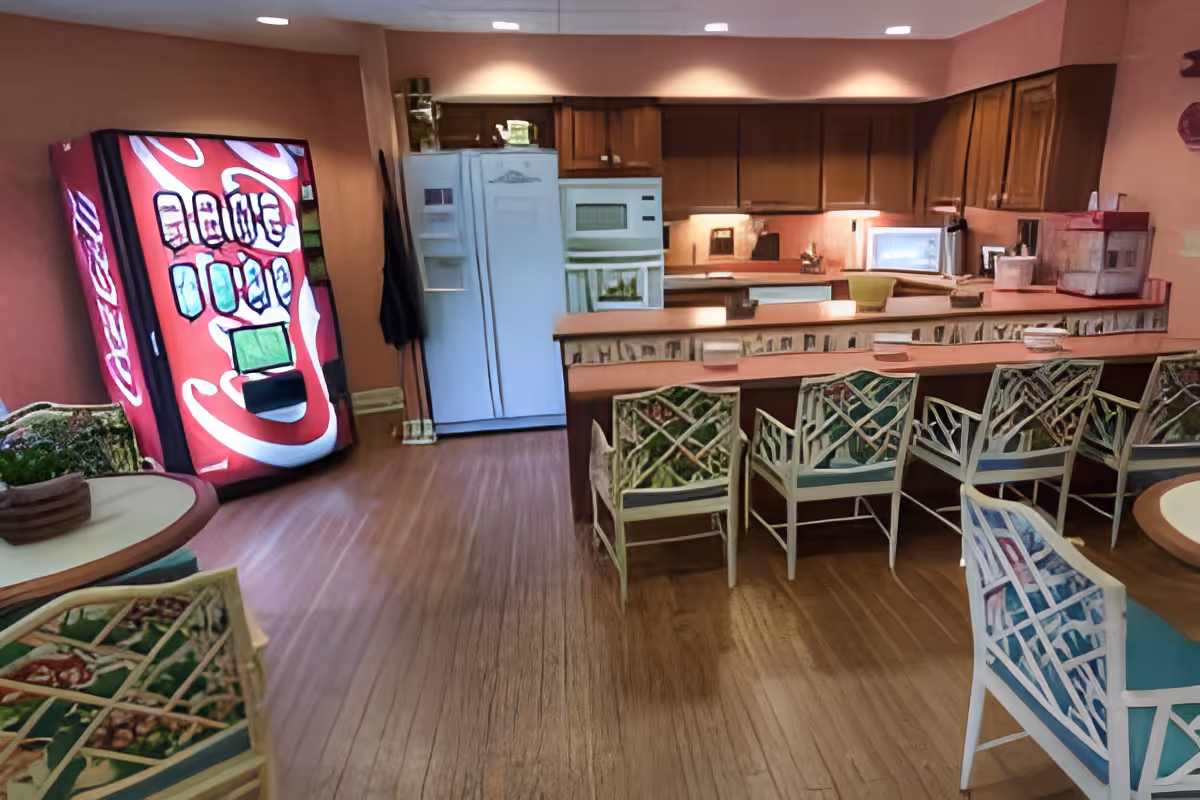 Interior view of a kitchen and dining area in a senior living facility. The room features wooden cabinets, a white refrigerator, microwave, and oven. There is a long counter with several chairs that have patterned cushions. To the left, there is a round table with chairs and a red Coca-Cola vending machine against the wall. The floor is wooden, and the walls are painted a warm color.
