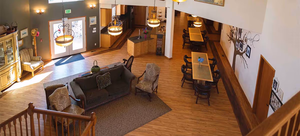 Interior view of a senior living facility common area with wooden flooring, a seating area with a sofa and armchairs on a rug, dining tables with chairs, decorative lighting fixtures, and a kitchen area in the background. The space is well-lit with natural light coming through glass doors.