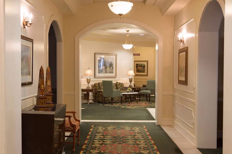 A carpeted hallway with an arched entry leading into a furnished sitting area with chairs, lamps, and framed artwork.