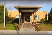 Front entrance of a low brick building with a covered walkway leading to double glass doors and surrounding landscaping.